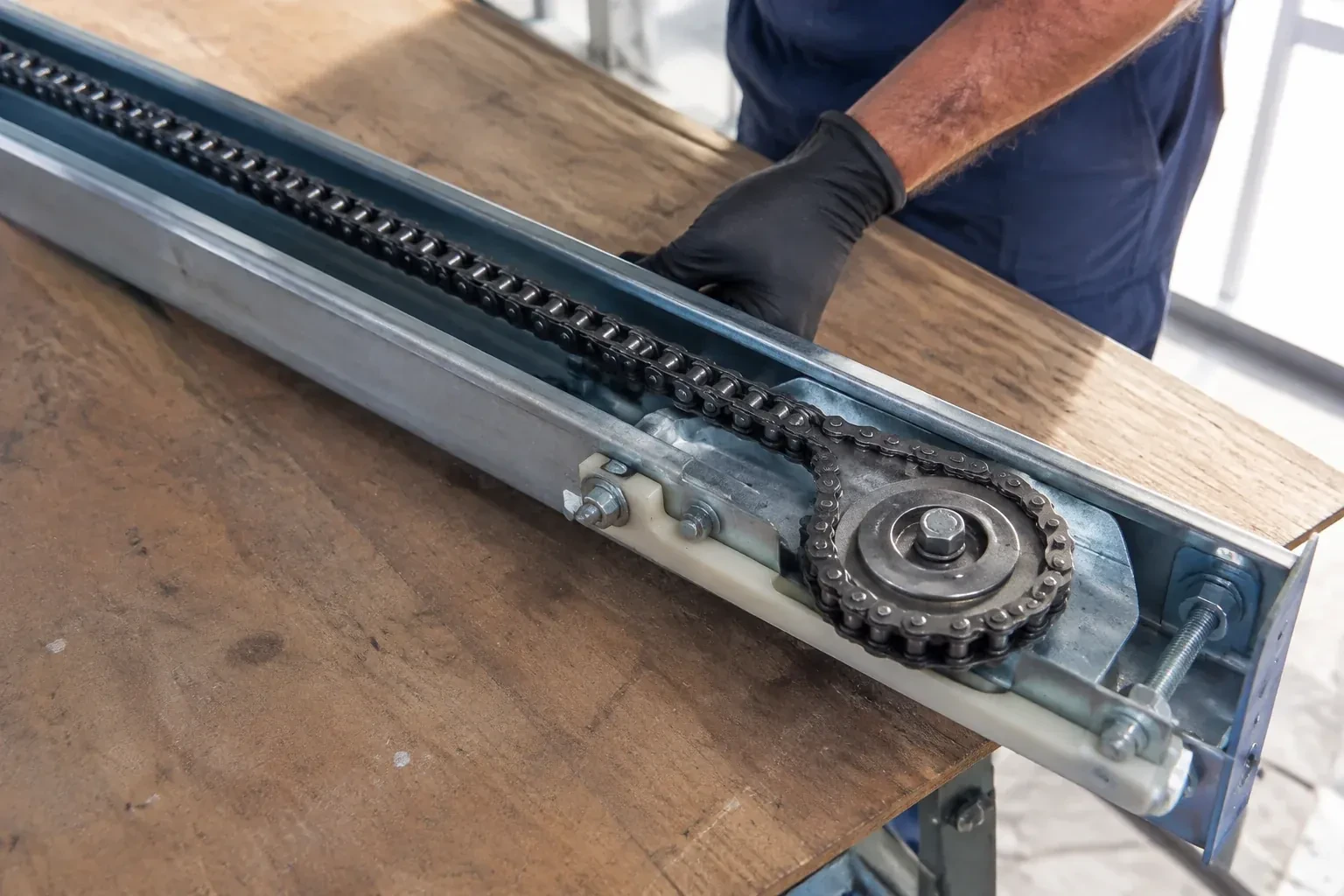 Gloved hand resting on a steel opener rail laid on a wooden bench, showing a drive chain looped around a sprocket wheel. — Chain-Drive Opener Repair, Wainfleet